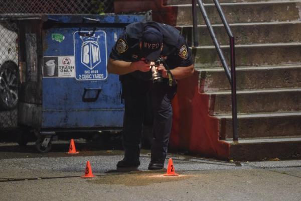 A police officer with a camera bends over to take a picture of the ground of the crime scene, wher<em></em>e there are are small pylons identifying evidence.
