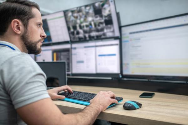 Attentive involved bearded young man with keyboard sitting watching in front of computer screens.