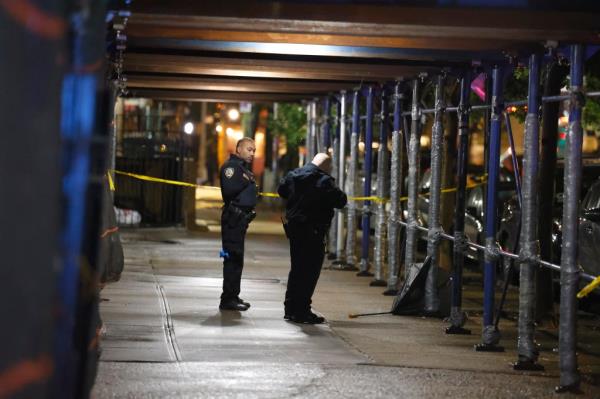 Two police officers stand underneath a scaffolding on a sidewalk cornered off by police tape. 