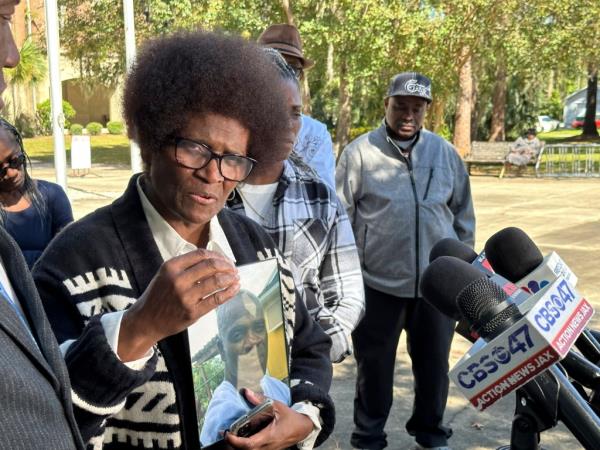 Mary Cure, the man's mother, speaks into news micropho<em></em>nes while she holds a photo of her son. 