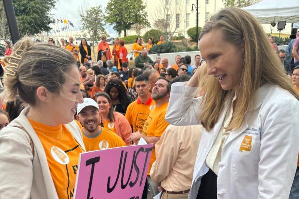 A woman in a white coat holding a sign while speaking with another person outside the Alabama Statehouse in Montgomery, Alabama.