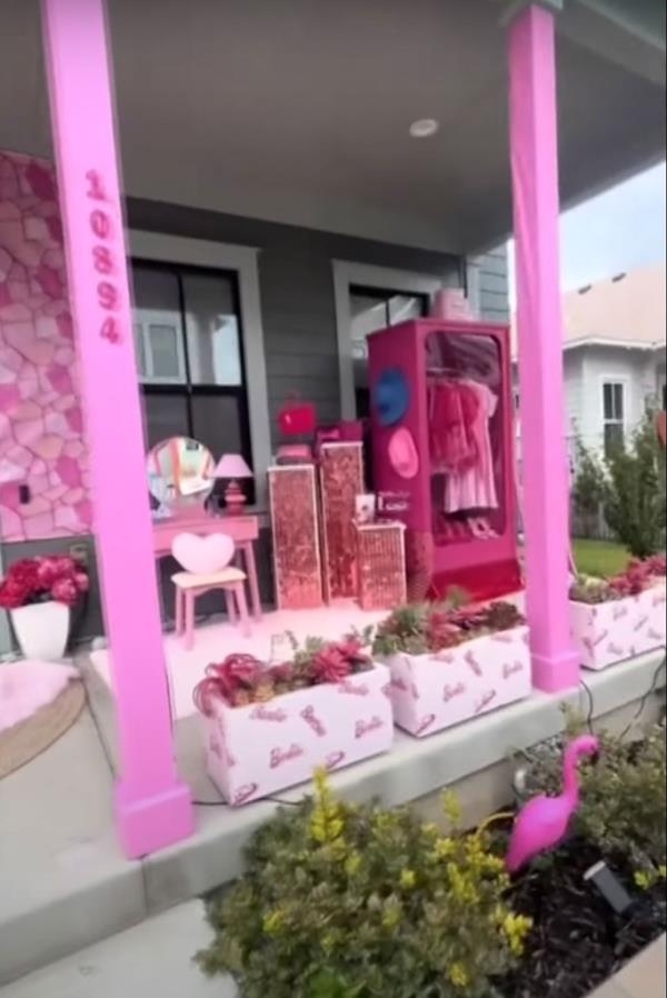 A home with multiple pink decorations on its front porch, including pink pillars and pink flower boxes with pink flowers, and a setup designed to look like Barbie's dressing room.