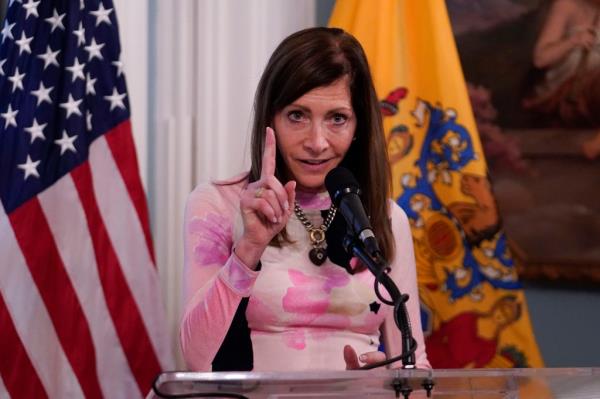 NJ First Lady Tammy Murphy gestures with one finger behind a microphone with the American and New Jersey flag behind her.