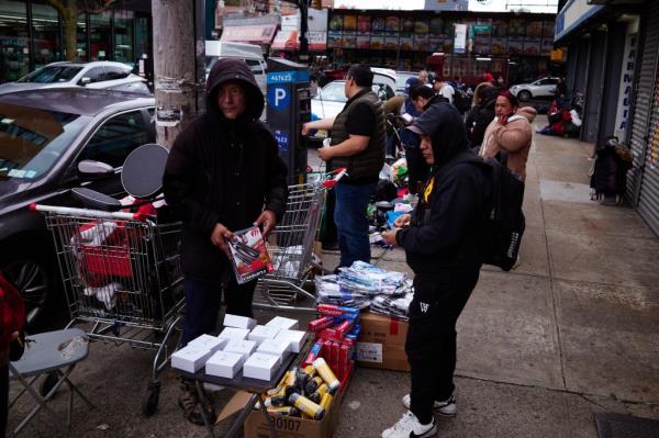 A group of people gathered around various goods sold on the sidewalk at the intersection of 91st St and Roosevelt Avenue, in an area known as Triangle 90 in Queens, New York