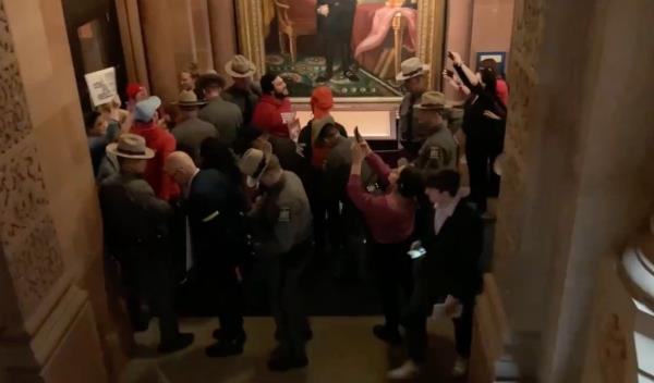 A group of housing activists being detained by troopers in a room with a painting, during a protest at Albany for a housing package.