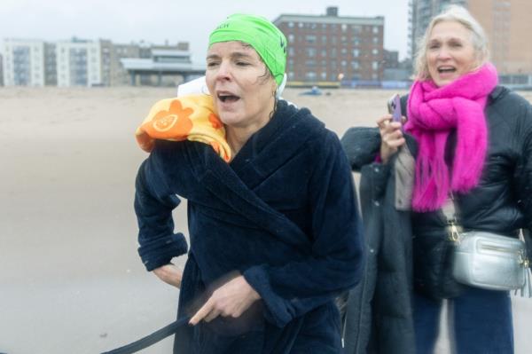 Wall Street Journal Editor-in-Chief Emma Tucker, wearing a bright green bathing cap, wraps a blue robe around her after swimming in the frigid water off Brighton Beach to call attention to detained reporter Evan Gershkovich's case. Behind her, a supporter wearing a bright pink scarf smiles.