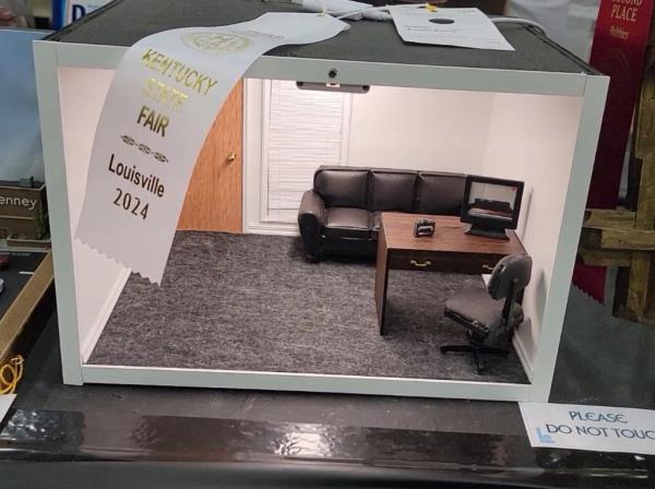 A miniature set with a couch, table, computer, camera, chair, window with blinds, and a door, and a state fair award ribbon draped over the top of the set.