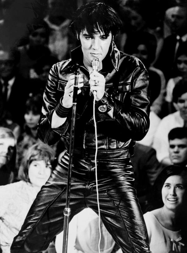 Elvis Presley in action during the NBC special. He's holding a microphone and is wearing a black leather outfit while he's singing. Audience members are visible in the background