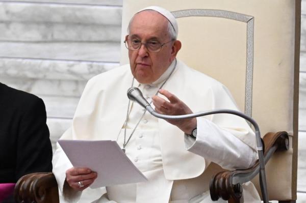 Pope Francis gestures with his left hand and holds papers in his right as he speaks into a microphone placed near his chair.