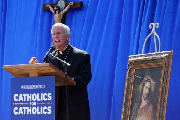 Bishop John Strickland speaks at a podium placed next to a portrait of Jesus. The podium has a sign attached that says 