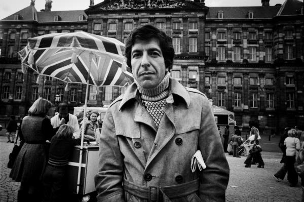 Leo<em></em>nard Cohen stands outside in from of a large building and food cart in the Netherlands. 