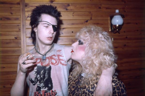 Sid Vicious, in eye patch, smokes a cigarette and puts his arm around Nancy Spunden. 