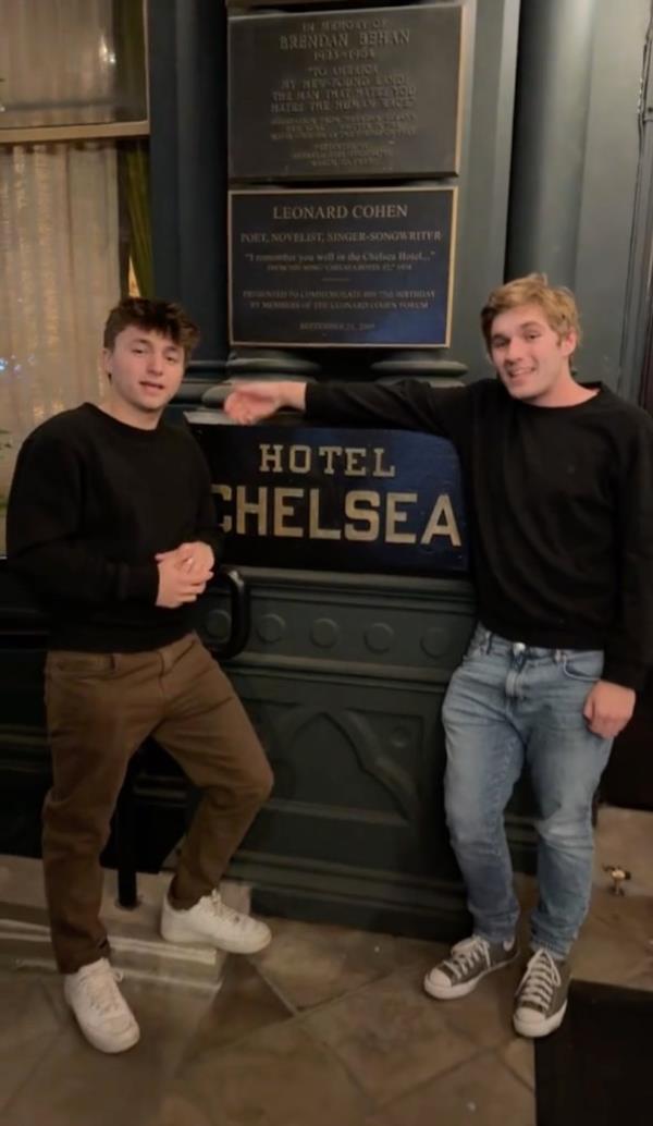 Two young men stand at the entrance to the Chelea Hotel, in front of a plaque that reads Hotel Chelsea. 