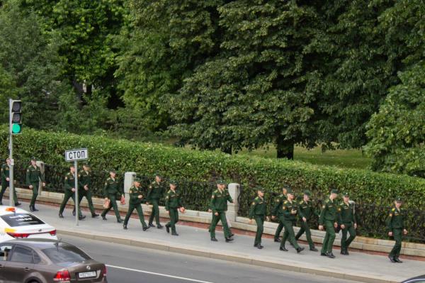 More than a dozen uniformed soldiers walk along a sidewalk in Moscow, with a park on one side of them and a roadway filled with cars on the other.