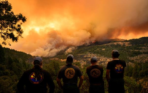 Four firefighters wearing t-shirts and ball caps stand shoulder to shoulder as they observe a forest fire moving through a valley in Butte County, California. The horizon is full of orange smoke and flames can been seen on the hill to the right.