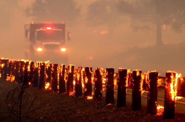 A fire engine drives through smoke-filled air past a flaming highway guard rail during the Park Fire in Tehama County, California.
