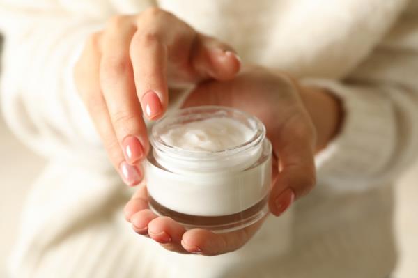 Woman in sweater showcasing her pink manicure while holding a jar of cosmetic cream