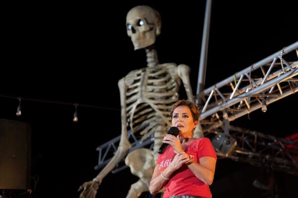 Arizona Republican US Senate candidate Kari Lake speaking into a microphone at a Halloween-themed rally, with a skeleton decoration in the background