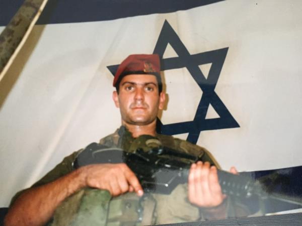 Aaron Cohen in his IDF military uniform holding a gun in front of an Israeli flag.