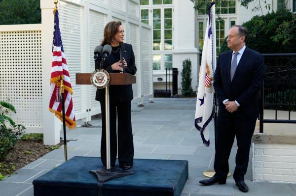 Vice President Kamala Harris speaking at a podium with micropho<em></em>nes and flags as Doug Emhoff looks on during a memorial tree planting ceremony, while protesters heckle from outside the gates.