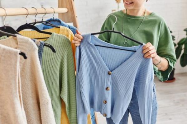 cropped view of tattooed woman with happy smile holding hanger with blue cardigan near rack with casual clothes at home, sustainable fashion and mindful co<em></em>nsumerism concept