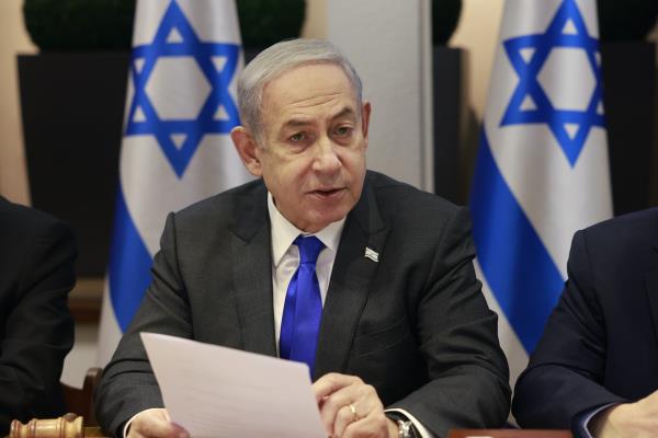 A man with grey hair in a suit talks in front of two Israeli flags